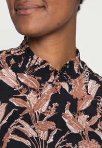 Haut noir avec un col smocké et un motif floral en nuances de brun et blanc. Présente des illustrations détaillées de plantes sur un tissu textur é.