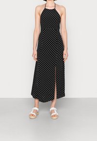 Schwarzes Halsbandkleid mit weißen Punkten, ausgestattet mit einem seitlichen Schlitz. Hergestellt aus leichtem Stoff mit einem fließenden Design.
