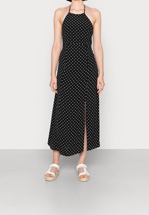 Robe noire à dos nu avec des pois blancs, dotée d'une fente sur le côté. Fabriquée en tissu léger avec un design fluide.