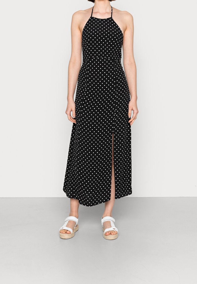 Schwarzes Halsbandkleid mit weißen Punkten, ausgestattet mit einem seitlichen Schlitz. Hergestellt aus leichtem Stoff mit einem fließenden Design.