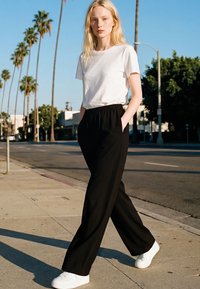 Ung kvinne med blondt hår går på solfylt fortau iført hvit t-skjorte, svarte vide bukser og hvite joggesko, med palmetrær i bakgrunnen.