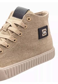 Zapatilla alta de lona beige con puntera redonda, suela beige y parche de logo negro. Presenta ojales circulares y cordones texturizados.