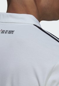 Gros plan d'une personne portant une chemise blanche à col avec des rayures noires et le texte "ARS AT HOME" au dos, près du col.