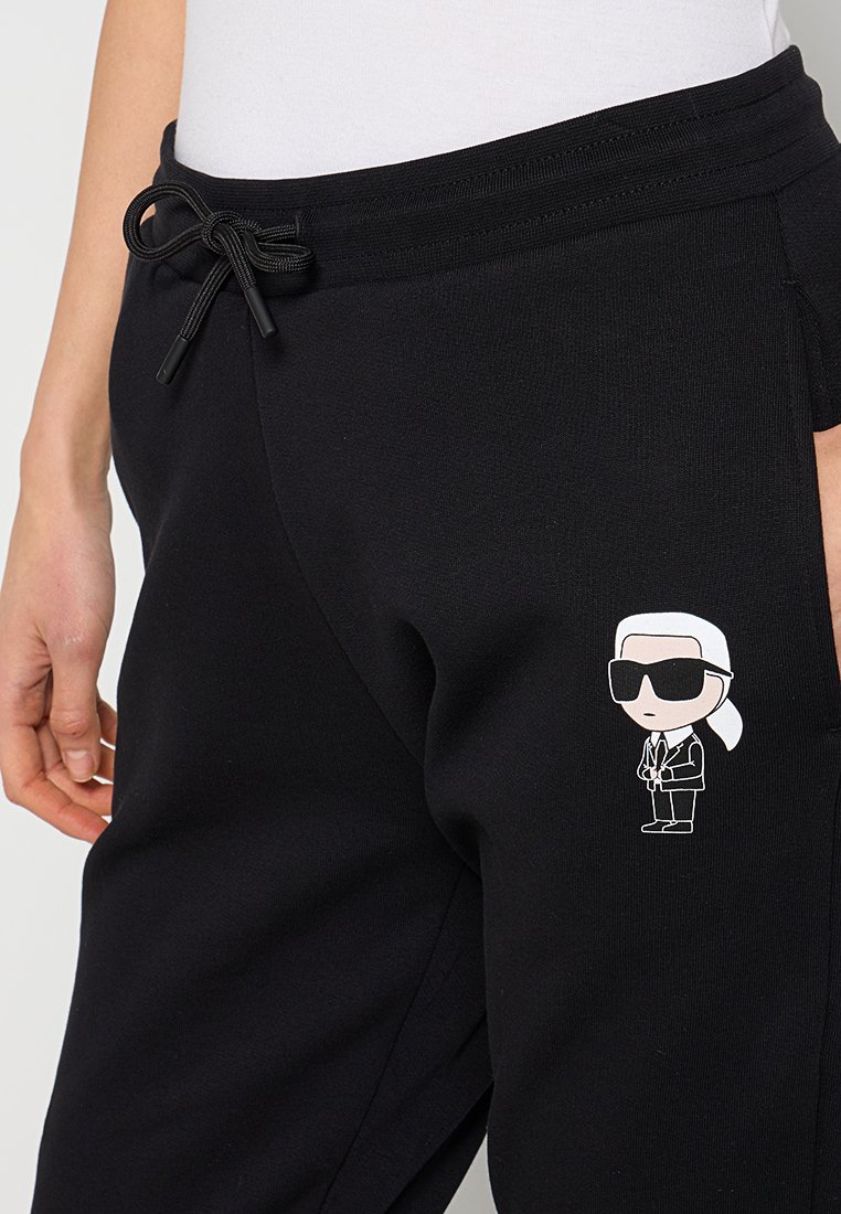 Un pantalon de survêtement noir en tissu doux, avec une taille à cordon de serrage et un graphisme de dessin animé sur la jambe gauche. Coupe standard et design décontracté.