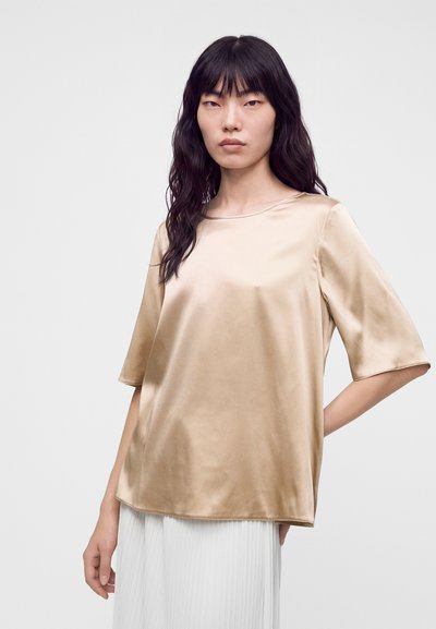 Femme aux longs cheveux foncés portant un top court en satin beige et une jupe blanche plissée, debout devant un fond clair uni.