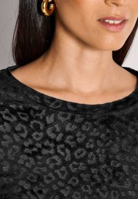 Vrouw draagt gouden hoepelringen en een zwart gestructureerd top met panterprint en een ronde nek.