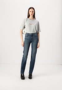 Grå t-skjorte med korte ermer og rød tekst, kombinert med tettsittende blå jeans og svarte sko, holder en hvit pose. Modellen står foran en ensfarget bakgrunn.