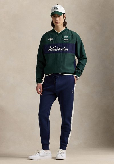 Grön och marinblå vindjacka med texten "Wimbledon", kombinerad med marinblå sweatpants med vita sidoränder och vita sneakers. Vit keps.