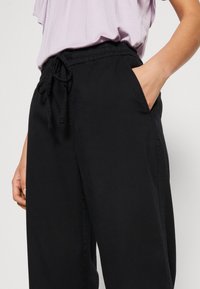 Pantalons noirs amples avec une taille élastique, détail de cordon de serrage et poches latérales, fabriqués en tissu doux.