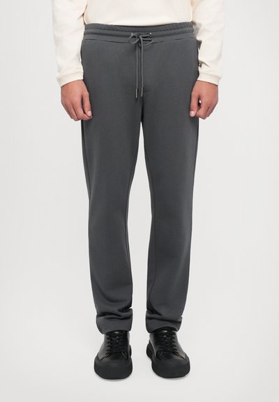 Pantalon de jogging gris avec une taille élastique et un cordon de serrage. Fabriqué en tissu doux, avec des poches latérales et des jambes effilées. Accompagné de baskets noires.