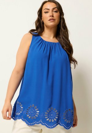 Kobieta o długich brązowych włosach, ubrana w niebieską, bezrękawnikową bluzkę z kwiatowym wycięciem przy dolnym brzegu, w połączeniu z jasnymi spodniami, stojąca na jednolitym tle.