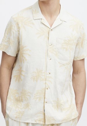 Homme portant une chemise beige clair à manches courtes avec boutons, à motif subtil de palmiers et poche poitrine.