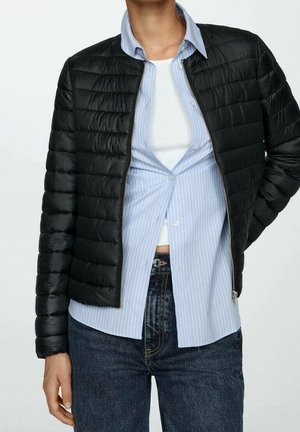 Persoon die een zwart doorgestikt jack draagt over een half dichtgeknoopte blauw gestreepte blouse en een wit hemd, gecombineerd met donkerblauwe jeans.