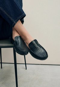 Zwarte loafers met een textuur van krokodillenpatroon, met een soepele bovenkant en gestikte details langs de zool, gedragen met donkere denim.