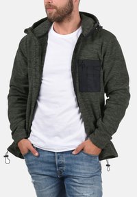 Chaqueta de forro polar verde oscuro con un bolsillo en el pecho con cremallera negra, capucha y dobladillos ajustables. Usada sobre una camiseta blanca y pantalones vaqueros azules.