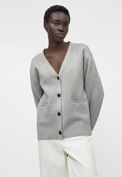 Γυναίκα που φοράει ένα ανοιχτό γκρι ζακέτα με κουμπιά μπροστά και εκρού παντελόνια με φαρδιές μπροστινές τσέπες, στέκεται μπροστά σε ένα απλό ανοιχτό φόντο.