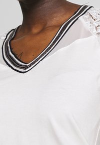 Biała bluzka z dekoltem w serek, z czarnymi i srebrnymi wykończeniami, delikatne koronkowe akcenty na rękawach, gładki materiał o lekki połysku.