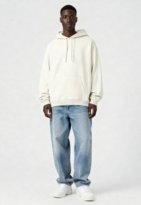 Jaunas vyras stovi, apsirengęs kreminės spalvos gobtuvu, laisvomis šviesiai mėlynomis džinsomis ir baltais sportbačiais prieš šviesią neutralios spalvos fono.