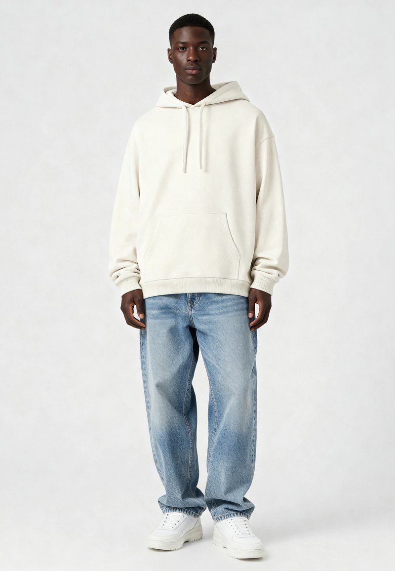 Jaunas vyras stovi, apsirengęs kreminės spalvos gobtuvu, laisvomis šviesiai mėlynomis džinsomis ir baltais sportbačiais prieš šviesią neutralios spalvos fono.