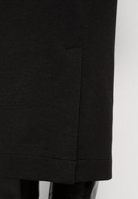 Gros plan de la partie inférieure d'une jupe noire avec une fente latérale, portée sur des bottes noires brillantes montant au genou.