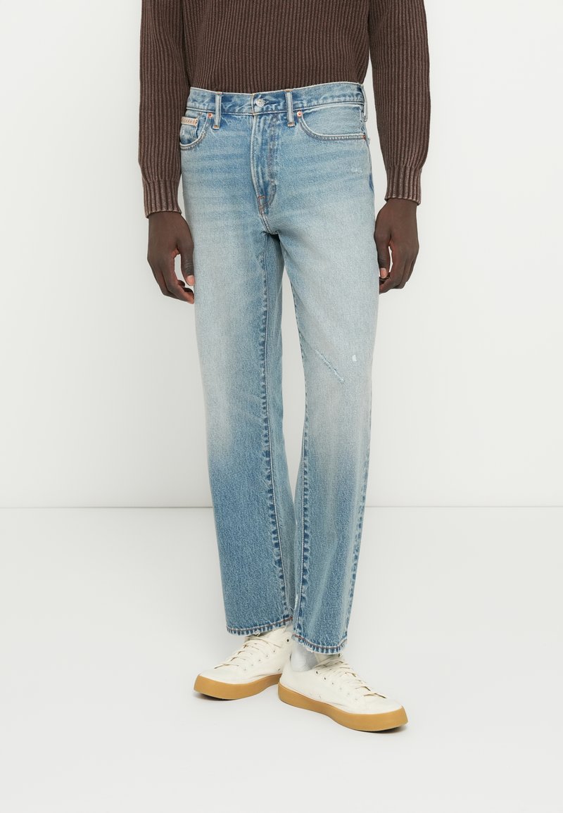 Ανοιχτό μπλε τζιν με ίσια γραμμή, με διακριτική ξεθώριαση και κλασικό σχέδιο με πέντε τσέπες. Συνδυασμένο με λευκά αθλητικά παπούτσια.