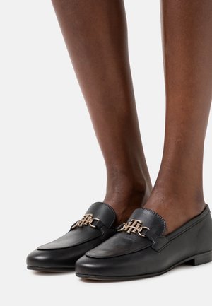 Czarne skórzane mokasyny z złotym łańcuszkiem, płaska podeszwa, okrągły nos i gładka tekstura, umieszczone na odkrytych nogach.