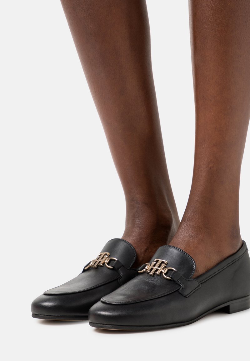 Zwarte leren instappers met een gouden kettingaccent, vlakke zool, ronde neus en een gladde textuur, gepositioneerd op blote benen.