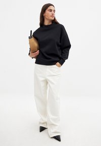 Sudadera negra de gran tamaño con un cuello alto, combinada con pantalones blancos de pierna ancha y zapatos negros de punta. Sosteniendo un bolso de mano marrón.