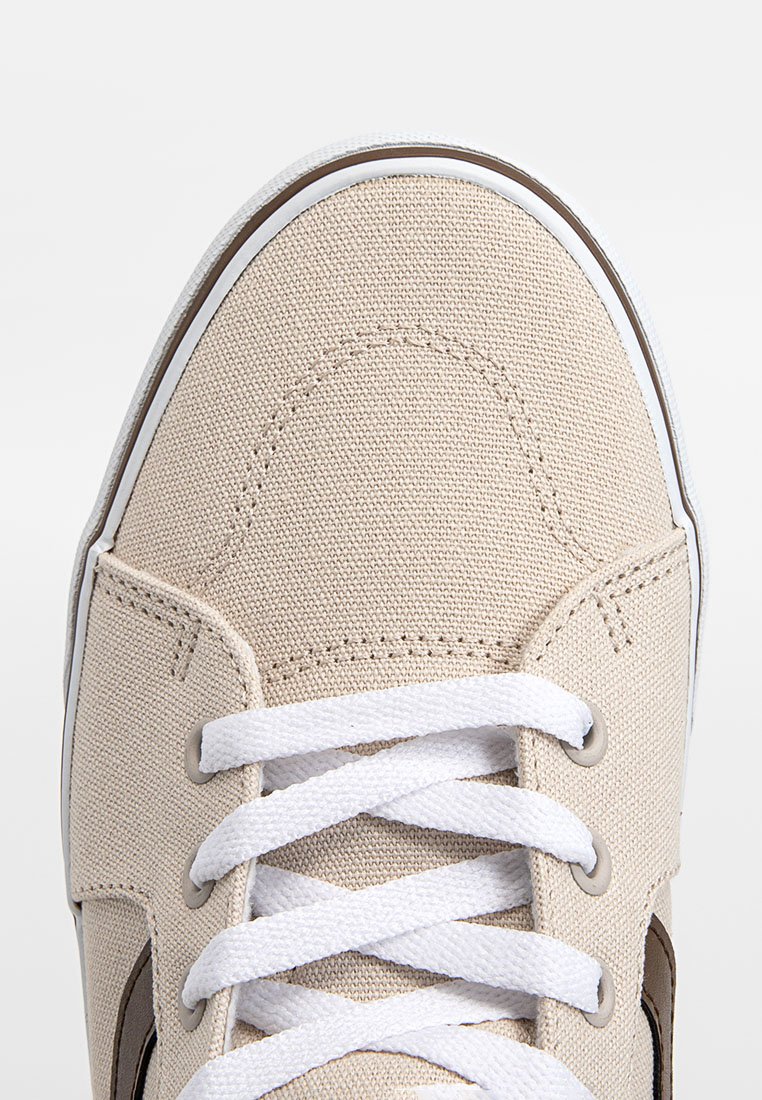 Zapatilla de lona beige con cordones blancos, que presenta un diseño limpio y una puntera redonda, acentuada por un logotipo oscuro en el lado.