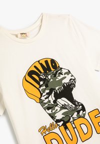 Vit bomulls T-shirt med ett grönt kamouflage-dinosaurietryck. Texten säger "Hello DINO DUDE" i fet gul och svart text.