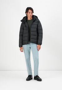 Sort pufferjakke med hætte, med en quiltet design og frontal lynlås, båret over en grøn sweater og lyseblå jeans. Sorte sko.