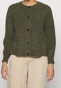 Olivgrön ullcardigan med rund halsringning, långa ärmar och sex stora svarta knappar framtill; ribbad fåll och manschetter.