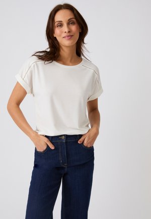 Donna con capelli castani fino alle spalle, in piedi con le mani nelle tasche, indossa una camicia bianca a maniche corte e jeans blu scuro su uno sfondo semplice.