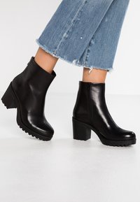 Stivaletti neri in pelle con tacco robusto, suola texturizzata e chiusura con zip. Presentano una finitura liscia e punta arrotondata.