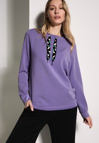 Felpa viola con maniche lunghe e scollatura tonda, caratterizzata da un motivo geometrico colorato sui lacci. Abbinata a pantaloni neri.
