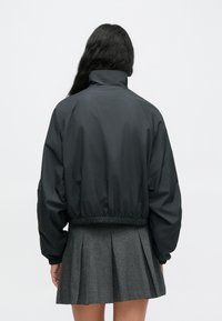 Μαύρο μπουφάν bomber με ψηλό γιακά και ελαστική μέση, συνδυασμένο με πλισέ γκρι φούστα, σε θέα από πίσω.