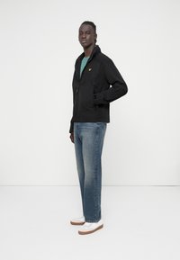 Black zip-up jacket with a textured finish, featuring a chest pocket and yellow logo. Paired with blue jeans and white sneakers with brown soles.