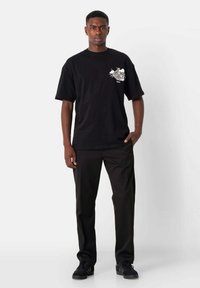 Camiseta de algodón negra con mangas cortas y estampado gráfico lateral; combinada con pantalones negros y zapatillas oscuras. Corte simple y relajado.