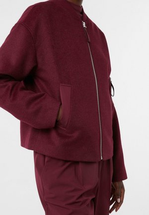 Persona che indossa una giacca rossa scura con zip e tasche laterali e pantaloni rossi scuri abbinati, in piedi davanti a uno sfondo bianco semplice.