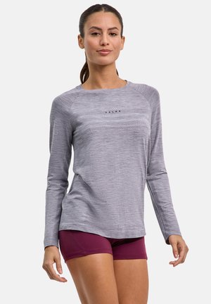 Vrouw draagt een lichtgrijs, langärmelig sportshirt en donkerpaarse shorts, staand met een zelfverzekerde houding tegen een effen achtergrond.