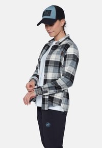 Camicia di flanella a quadri bianco e nero con polsini con bottoni, indossata sopra una maglietta leggera. Abbinata a un cappellino blu e nero con un logo.