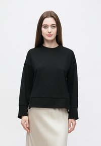 Μαύρο φούτερ με μακριά μανίκια, κατασκευασμένο από απαλό ύφασμα, με ελαφρώς κατεβατό ώμο και ευθεία τελική ραφ�ή με φερμουάρ στα πλαϊνά.