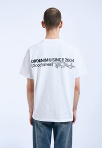 Bílý bavlněný tričko s černým textem na zadní straně: "DRDENIM© OD ROKU 2004 (Dobré časy)" s graffiti-style akcentem. Volný střih.