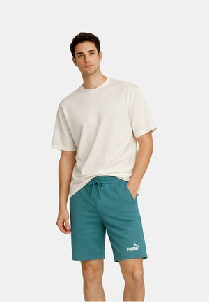 Giovane uomo che indossa una maglietta bianca larga e pantaloncini turchesi Puma con coulisse, in piedi con una mano in tasca davanti a uno sfondo semplice.