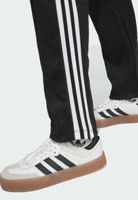 Bílé a černé tenisky Adidas se třemi černými pruhy, gumenou podrážkou, nošené s černými kalhotami se třemi bílými pruhy po stranách.