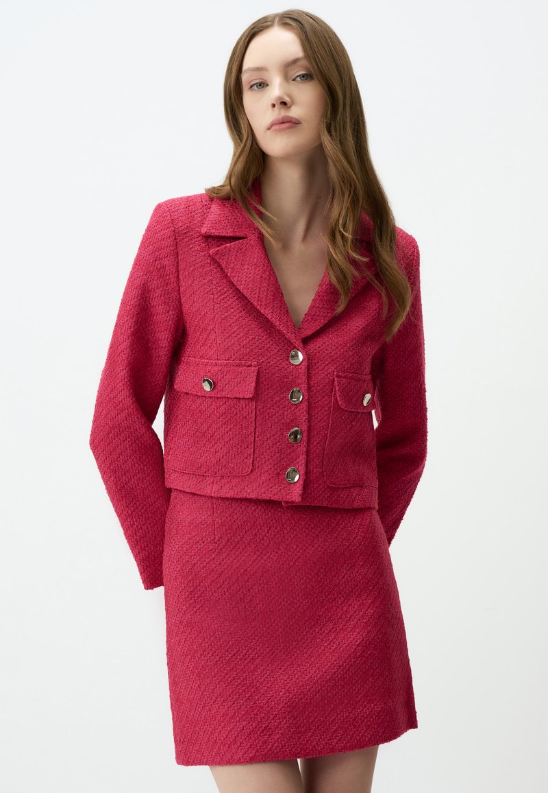 Blazer texturé rose vif avec un col, quatre poches et des boutons en argent, associé à une jupe mini texturée assortie.