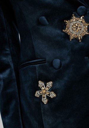 Prenda de terciopelo azul marino que presenta dos broches ornamentales: un diseño circular en oro y gemas, y un broche en forma de estrella con acentos brillantes.