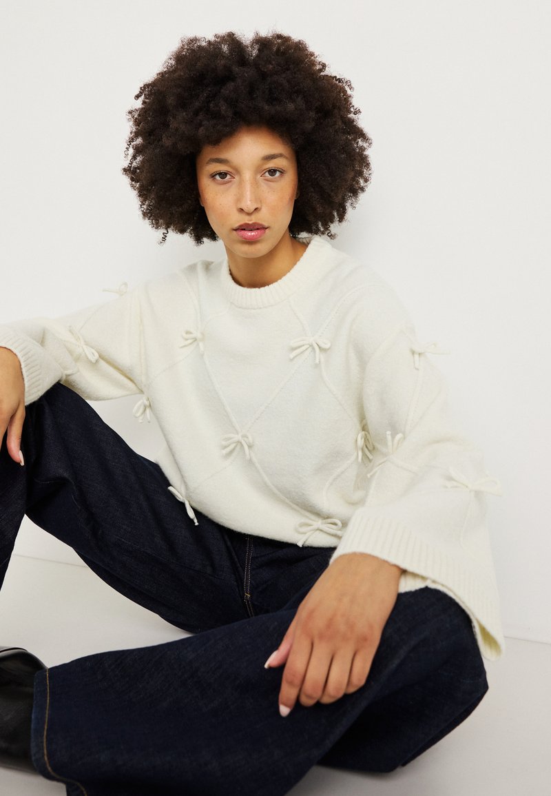 Balta megzta sveteris su kaspinų akcentais, raukšlėmis rankovėse ir laisvu siluetu, derinamas su tamsiais džinsiniais kelnėmis. Išskirtinė ir kruopščiai sukurta dizaino struktūra.