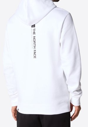 Mann trägt einen weißen Hoodie mit einem vertikalen schwarzen "The North Face"-Logo und Schriftzug auf dem Rücken, kombiniert mit schwarzen Hosen.