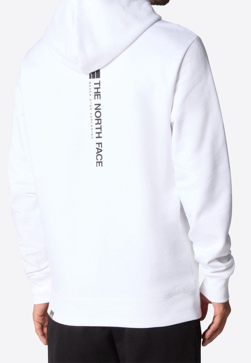Homme portant un sweat à capuche blanc avec un logo vertical noir « The North Face » et un texte dans le dos, associé à un pantalon noir.
